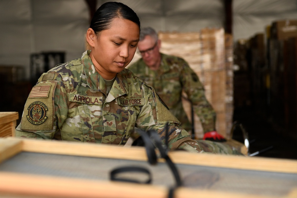 A U.S. Air Force service member named Arriola is working in what appears to be a warehouse or storage facility, handling wooden crates or equipment. A second service member is visible in the background wearing red gloves and working with palletized materials. Both personnel are wearing OCP (Operational Camouflage Pattern) uniforms.