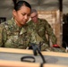 A U.S. Air Force service member named Arriola is working in what appears to be a warehouse or storage facility, handling wooden crates or equipment. A second service member is visible in the background wearing red gloves and working with palletized materials. Both personnel are wearing OCP (Operational Camouflage Pattern) uniforms.