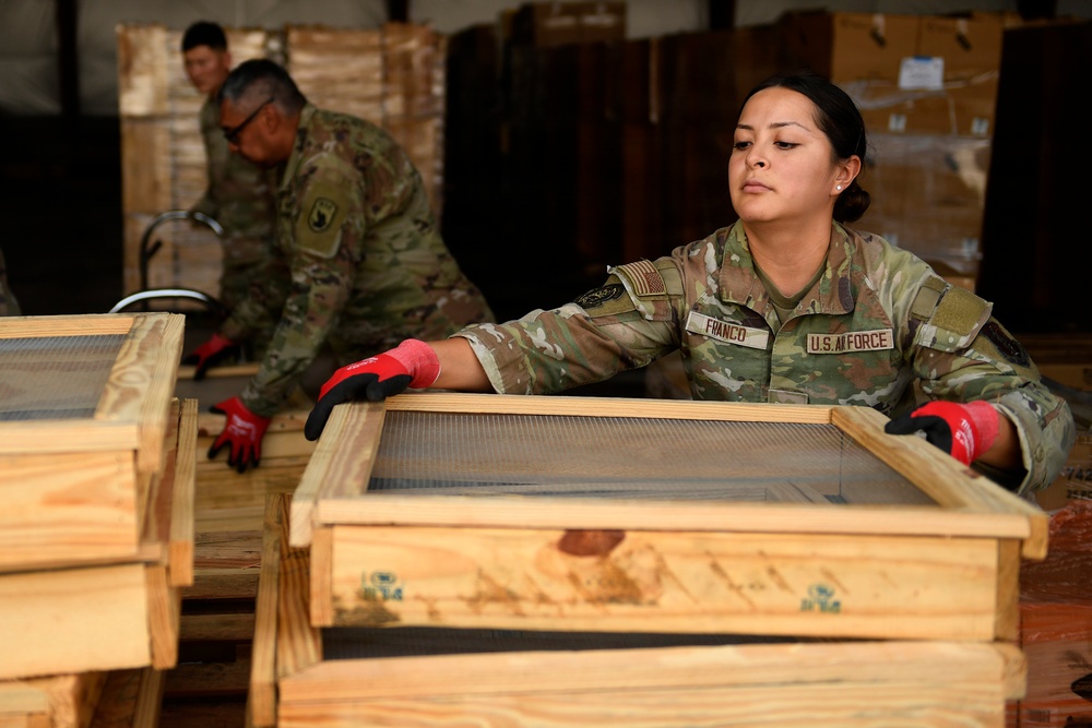 A U.S. Air Force service member named Franco is working in what appears to be a warehouse or logistics facility, handling wooden crates or frames with a mesh/screen insert. She is wearing OCP (Operational Camouflage Pattern) uniform and red-and-black work gloves. Two additional military personnel in the background are engaged in similar logistics or cargo handling activities.