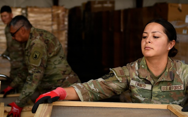 A U.S. Air Force service member named Franco is working in what appears to be a warehouse or logistics facility, handling wooden crates or frames with a mesh/screen insert. She is wearing OCP (Operational Camouflage Pattern) uniform and red-and-black work gloves. Two additional military personnel in the background are engaged in similar logistics or cargo handling activities.