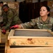 A U.S. Air Force service member named Franco is working in what appears to be a warehouse or logistics facility, handling wooden crates or frames with a mesh/screen insert. She is wearing OCP (Operational Camouflage Pattern) uniform and red-and-black work gloves. Two additional military personnel in the background are engaged in similar logistics or cargo handling activities.