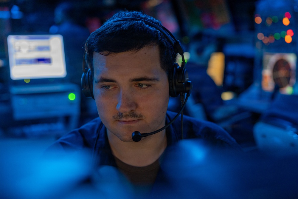 A young man wearing a communications headset is focused on screens in a dimly lit, blue-toned operations environment. Multiple monitors and equipment are visible in the background, suggesting a command center, military operations room, or broadcast control setting. The lighting is predominantly blue with accent colors from various screens and indicator lights.