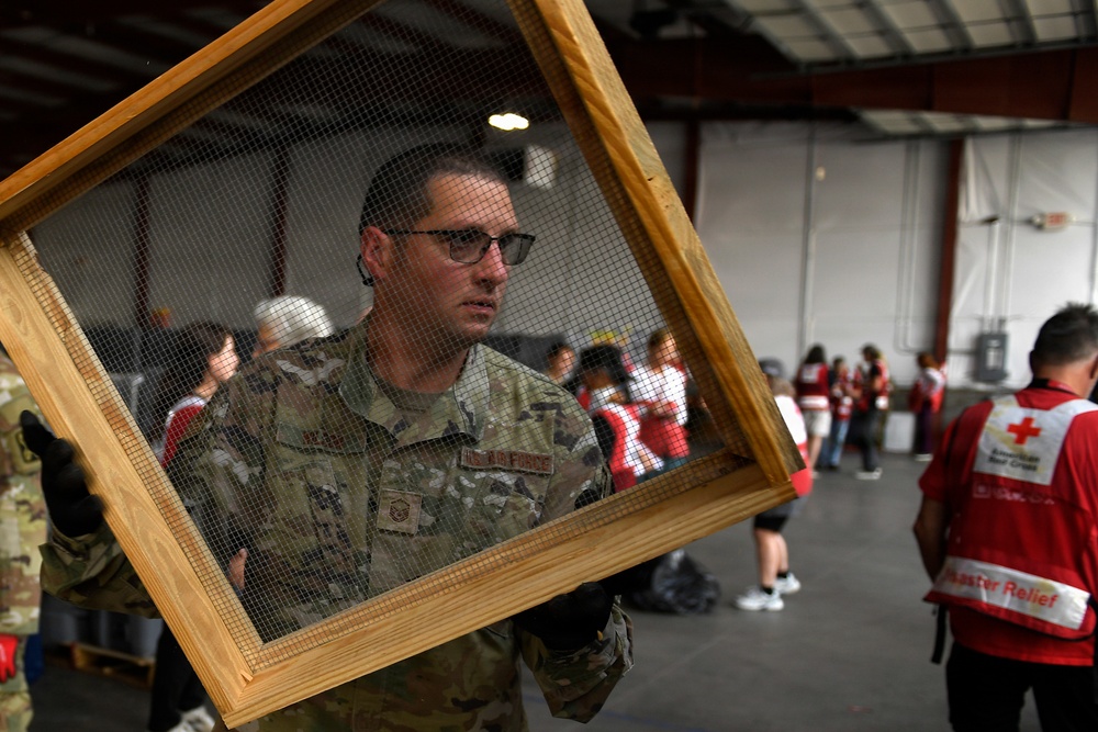 A U.S. Air Force service member in camouflage uniform carries a wooden-framed wire mesh screen inside what appears to be a disaster relief staging area. In the background, multiple American Red Cross volunteers in red vests marked 'Disaster Relief' are gathered, suggesting a coordinated emergency response operation. The scene depicts military and civilian humanitarian organizations working together.