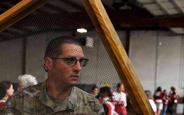 A U.S. Air Force service member in camouflage uniform carries a wooden-framed wire mesh screen inside what appears to be a disaster relief staging area. In the background, multiple American Red Cross volunteers in red vests marked 'Disaster Relief' are gathered, suggesting a coordinated emergency response operation. The scene depicts military and civilian humanitarian organizations working together.
