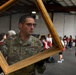 A U.S. Air Force service member in camouflage uniform carries a wooden-framed wire mesh screen inside what appears to be a disaster relief staging area. In the background, multiple American Red Cross volunteers in red vests marked 'Disaster Relief' are gathered, suggesting a coordinated emergency response operation. The scene depicts military and civilian humanitarian organizations working together.