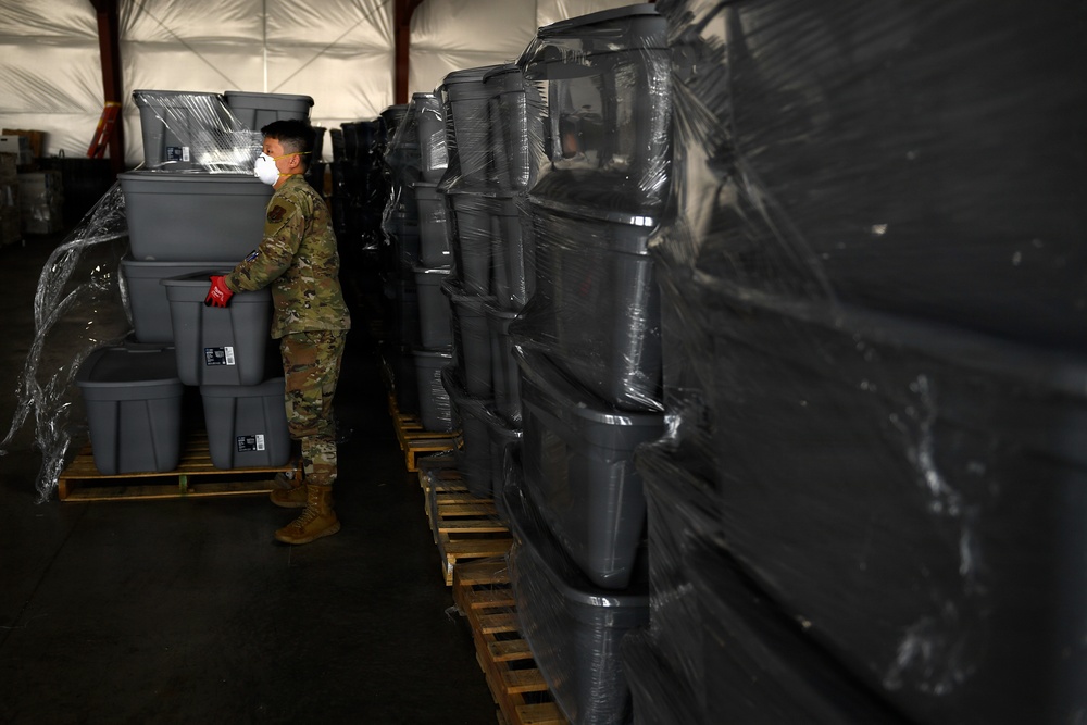 A military service member wearing camouflage uniform and an N95/respirator face mask is working inside a large tent or temporary warehouse structure. The individual is handling gray plastic storage bins/totes that are stacked on wooden pallets, some wrapped in clear plastic shrink wrap. The scene suggests a large-scale logistics or supply distribution operation.
