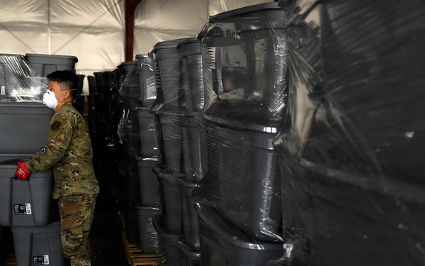 A military service member wearing camouflage uniform and an N95/respirator face mask is working inside a large tent or temporary warehouse structure. The individual is handling gray plastic storage bins/totes that are stacked on wooden pallets, some wrapped in clear plastic shrink wrap. The scene suggests a large-scale logistics or supply distribution operation.