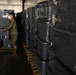 A military service member wearing camouflage uniform and an N95/respirator face mask is working inside a large tent or temporary warehouse structure. The individual is handling gray plastic storage bins/totes that are stacked on wooden pallets, some wrapped in clear plastic shrink wrap. The scene suggests a large-scale logistics or supply distribution operation.