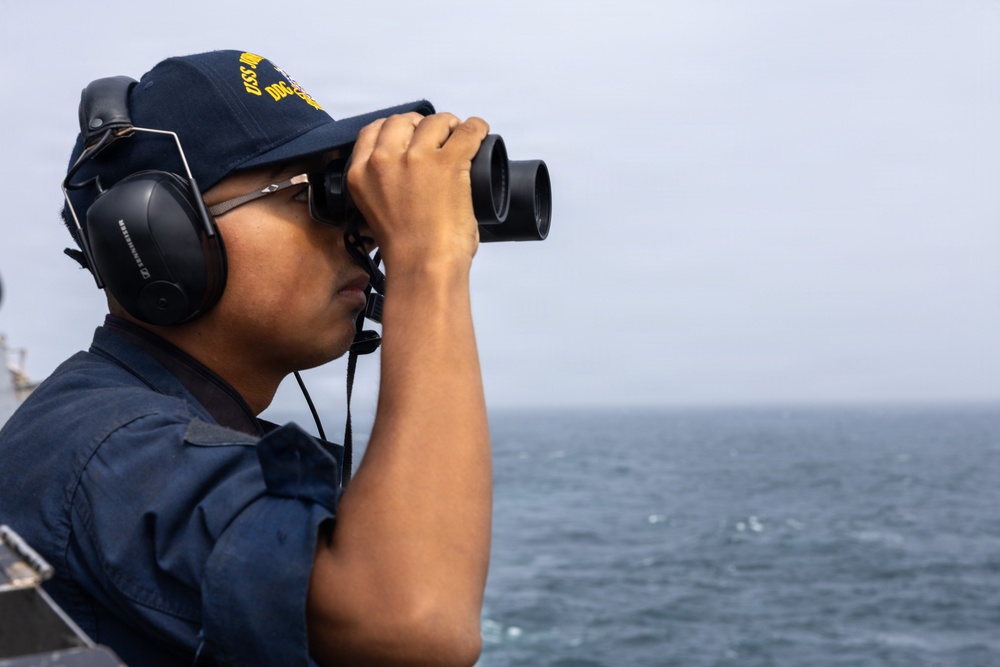 A U.S. Navy sailor is standing watch on the deck of a naval vessel, scanning the horizon with large binoculars. The sailor is wearing standard Navy working uniform (NWU), a Navy ball cap with 'USS' and 'DDG' visible on it, and Sennheiser communication headphones. The background shows open ocean under an overcast sky.