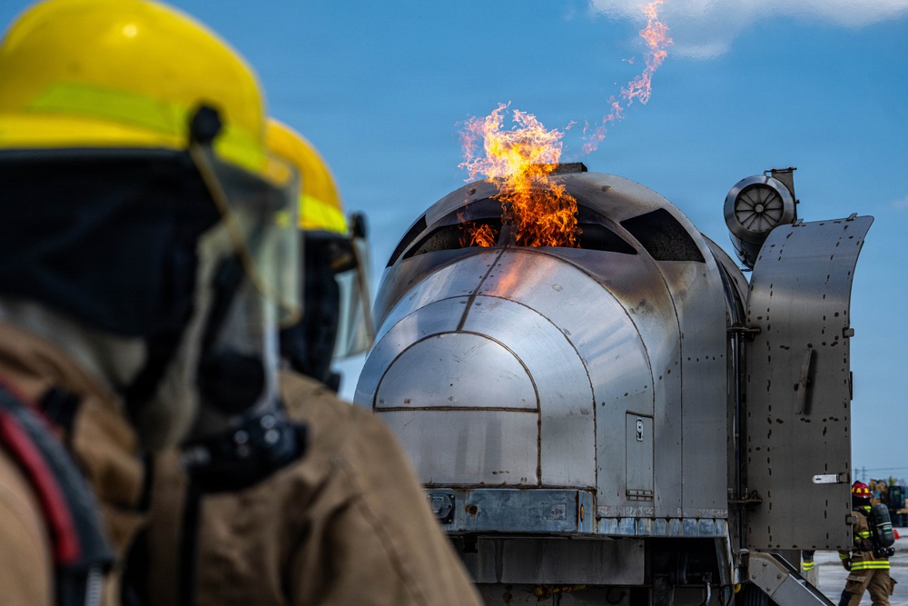 A firefighter in full protective gear including a yellow helmet and breathing apparatus observes a controlled fire training exercise. A mock aircraft fuselage or cockpit section simulator is actively burning, with flames visible at the top of the structure. Additional firefighters in turnout gear are visible in the background, indicating this is a coordinated training drill.