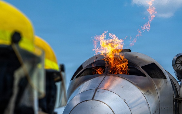 A firefighter in full protective gear including a yellow helmet and breathing apparatus observes a controlled fire training exercise. A mock aircraft fuselage or cockpit section simulator is actively burning, with flames visible at the top of the structure. Additional firefighters in turnout gear are visible in the background, indicating this is a coordinated training drill.