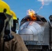 A firefighter in full protective gear including a yellow helmet and breathing apparatus observes a controlled fire training exercise. A mock aircraft fuselage or cockpit section simulator is actively burning, with flames visible at the top of the structure. Additional firefighters in turnout gear are visible in the background, indicating this is a coordinated training drill.