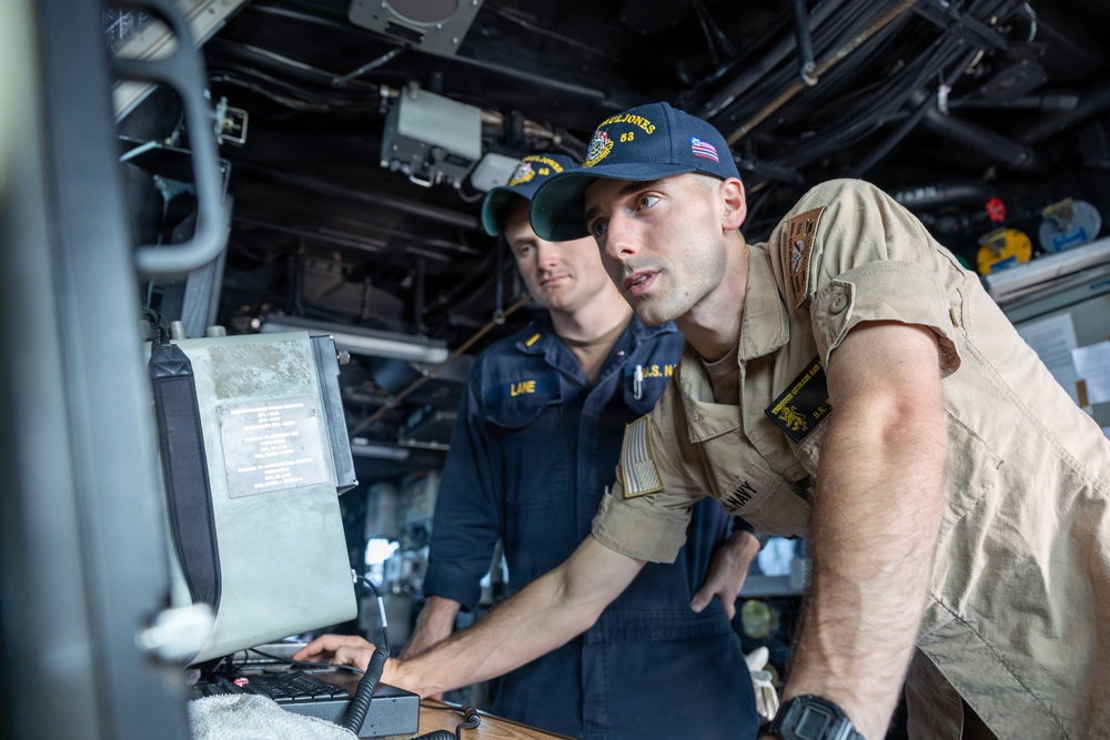 Two U.S. Navy personnel are working at a console aboard a naval vessel, likely on the bridge or combat information center. The sailor in the foreground, wearing khaki utilities and a cap reading 'Paul Jones 53,' is leaning over a monitor while the sailor behind him, wearing navy blue coveralls with a name tape reading 'LANE,' observes. Both appear to be engaged in navigation or operational duties.