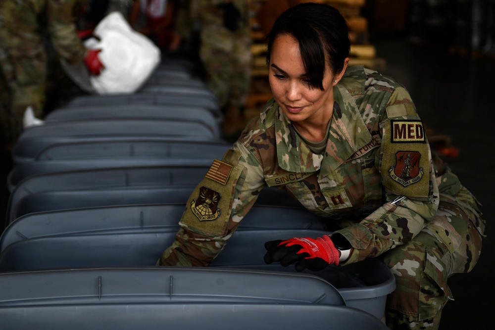 A female U.S. Air Force National Guard medical officer with the last name 'Sinclair' is working with gray plastic storage containers or bins in what appears to be a warehouse or logistics facility. She is wearing OCP (Operational Camouflage Pattern) uniform with a 'MED' tab, captain's rank insignia, and red work gloves. Other personnel in similar uniforms are visible in the background performing similar tasks.