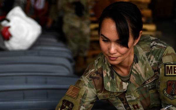 A female U.S. Air Force National Guard medical officer with the last name 'Sinclair' is working with gray plastic storage containers or bins in what appears to be a warehouse or logistics facility. She is wearing OCP (Operational Camouflage Pattern) uniform with a 'MED' tab, captain's rank insignia, and red work gloves. Other personnel in similar uniforms are visible in the background performing similar tasks.