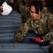 A female U.S. Air Force National Guard medical officer with the last name 'Sinclair' is working with gray plastic storage containers or bins in what appears to be a warehouse or logistics facility. She is wearing OCP (Operational Camouflage Pattern) uniform with a 'MED' tab, captain's rank insignia, and red work gloves. Other personnel in similar uniforms are visible in the background performing similar tasks.