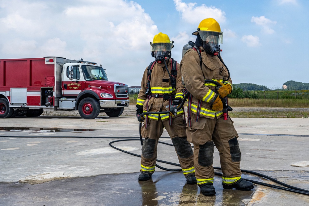 Two United States Air Force firefighters in full personal protective equipment stand on a wet concrete surface, likely during a training exercise. They are wearing tan turnout gear with yellow reflective striping, yellow helmets, and full-face SCBA (Self-Contained Breathing Apparatus) masks. A red and white USAF fire truck is visible in the background.