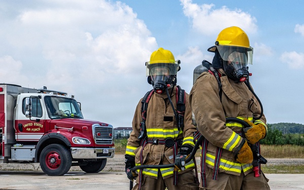 Two United States Air Force firefighters in full personal protective equipment stand on a wet concrete surface, likely during a training exercise. They are wearing tan turnout gear with yellow reflective striping, yellow helmets, and full-face SCBA (Self-Contained Breathing Apparatus) masks. A red and white USAF fire truck is visible in the background.