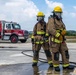 Two United States Air Force firefighters in full personal protective equipment stand on a wet concrete surface, likely during a training exercise. They are wearing tan turnout gear with yellow reflective striping, yellow helmets, and full-face SCBA (Self-Contained Breathing Apparatus) masks. A red and white USAF fire truck is visible in the background.