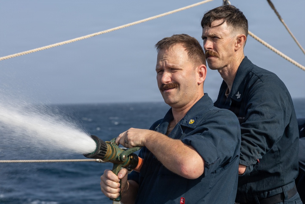 Two naval personnel in dark blue uniforms are conducting what appears to be a firefighting drill or washdown exercise aboard a vessel at sea. The foreground sailor is actively operating a high-pressure fire hose nozzle, while the second sailor observes from behind. The open ocean is visible in the background, confirming an at-sea operational setting.