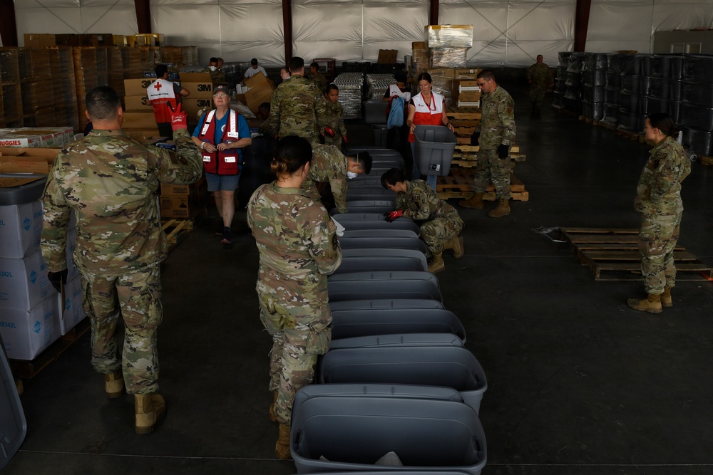 Military personnel in camouflage uniforms and American Red Cross volunteers are working together in a large warehouse facility, assembling or organizing disaster relief supplies. A row of gray plastic storage bins/totes is lined up in the foreground, while pallets of boxed goods, water, and other supplies are stacked in the background. This appears to be a disaster relief staging and distribution operation.