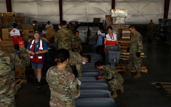 Military personnel in camouflage uniforms and American Red Cross volunteers are working together in a large warehouse facility, assembling or organizing disaster relief supplies. A row of gray plastic storage bins/totes is lined up in the foreground, while pallets of boxed goods, water, and other supplies are stacked in the background. This appears to be a disaster relief staging and distribution operation.