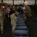 Military personnel in camouflage uniforms and American Red Cross volunteers are working together in a large warehouse facility, assembling or organizing disaster relief supplies. A row of gray plastic storage bins/totes is lined up in the foreground, while pallets of boxed goods, water, and other supplies are stacked in the background. This appears to be a disaster relief staging and distribution operation.