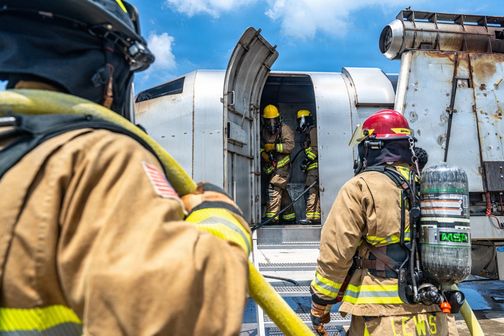 Firefighters in full personal protective equipment (PPE) are conducting what appears to be an aircraft firefighting training exercise involving a decommissioned or mock aircraft fuselage. Two firefighters are visible outside the aircraft while two others are positioned inside the open door, with a yellow fire hose being fed into the structure. The aircraft shows signs of heavy use and weathering consistent with a training prop.