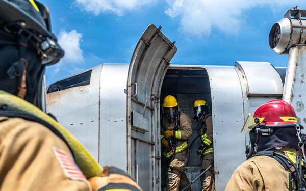 Firefighters in full personal protective equipment (PPE) are conducting what appears to be an aircraft firefighting training exercise involving a decommissioned or mock aircraft fuselage. Two firefighters are visible outside the aircraft while two others are positioned inside the open door, with a yellow fire hose being fed into the structure. The aircraft shows signs of heavy use and weathering consistent with a training prop.