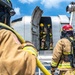 Firefighters in full personal protective equipment (PPE) are conducting what appears to be an aircraft firefighting training exercise involving a decommissioned or mock aircraft fuselage. Two firefighters are visible outside the aircraft while two others are positioned inside the open door, with a yellow fire hose being fed into the structure. The aircraft shows signs of heavy use and weathering consistent with a training prop.