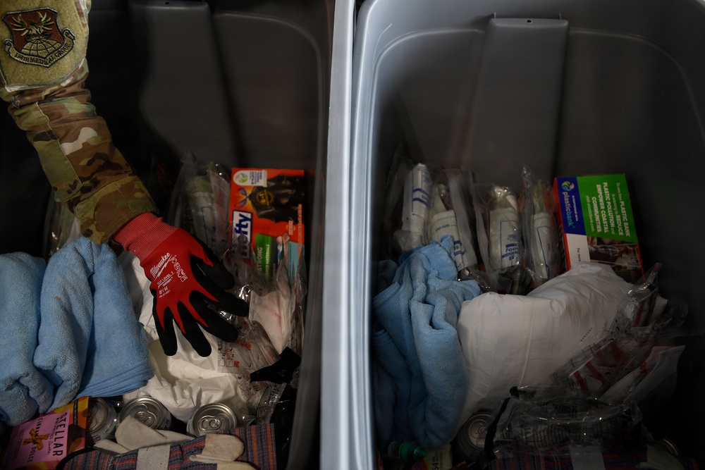 The image shows two gray plastic storage bins or totes filled with various supplies, including work gloves, towels, packaged items, and canned goods. A person wearing military camouflage (multicam pattern) clothing with a unit patch is visible at the upper left, reaching into or near the left bin. The contents suggest these may be emergency, relief, or disaster response supply kits.