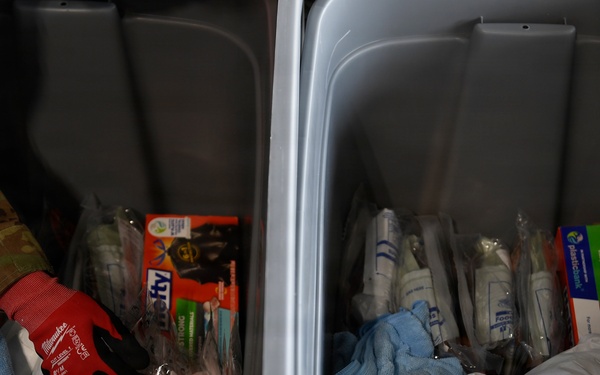 The image shows two gray plastic storage bins or totes filled with various supplies, including work gloves, towels, packaged items, and canned goods. A person wearing military camouflage (multicam pattern) clothing with a unit patch is visible at the upper left, reaching into or near the left bin. The contents suggest these may be emergency, relief, or disaster response supply kits.