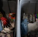 The image shows two gray plastic storage bins or totes filled with various supplies, including work gloves, towels, packaged items, and canned goods. A person wearing military camouflage (multicam pattern) clothing with a unit patch is visible at the upper left, reaching into or near the left bin. The contents suggest these may be emergency, relief, or disaster response supply kits.