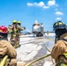 A group of firefighters in full protective gear and SCBA (Self-Contained Breathing Apparatus) are conducting a live fire training exercise on what appears to be an airfield or tarmac. They are directing water hoses at a silver aircraft fuselage mockup that has an active flame burning on top. The scene is set under a clear blue sky with scattered clouds during daytime.