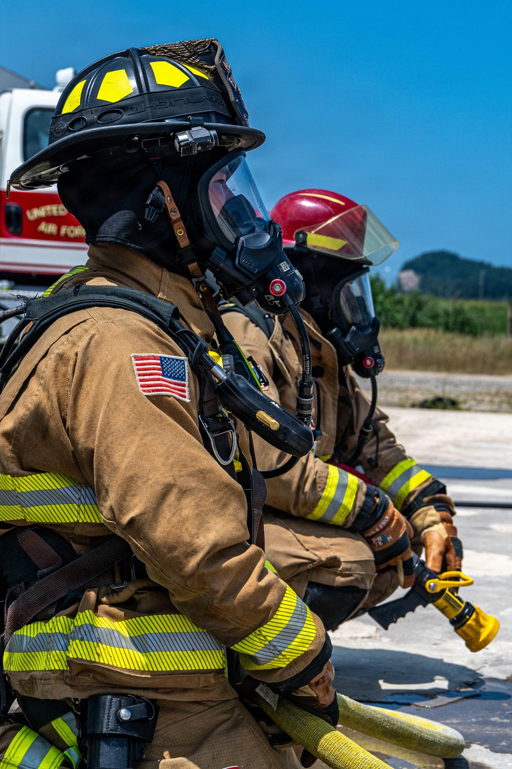 Two firefighters in full personal protective equipment are positioned side by side, holding a fire hose during what appears to be a training exercise or active firefighting operation. The foreground firefighter wears a black helmet with yellow accents and displays a US flag patch on the sleeve, with a vehicle partially visible bearing 'United States Air Force' markings. Both personnel are equipped with SCBA (Self-Contained Breathing Apparatus) masks and full turnout gear.