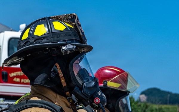 Two firefighters in full personal protective equipment are positioned side by side, holding a fire hose during what appears to be a training exercise or active firefighting operation. The foreground firefighter wears a black helmet with yellow accents and displays a US flag patch on the sleeve, with a vehicle partially visible bearing 'United States Air Force' markings. Both personnel are equipped with SCBA (Self-Contained Breathing Apparatus) masks and full turnout gear.