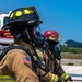 Two firefighters in full personal protective equipment are positioned side by side, holding a fire hose during what appears to be a training exercise or active firefighting operation. The foreground firefighter wears a black helmet with yellow accents and displays a US flag patch on the sleeve, with a vehicle partially visible bearing 'United States Air Force' markings. Both personnel are equipped with SCBA (Self-Contained Breathing Apparatus) masks and full turnout gear.