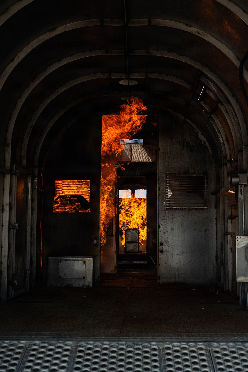 The image shows the interior of a metal-walled corridor or tunnel structure with an arched ceiling, through which intense open flames are visible in the middle and background sections. The fire appears to be burning actively through a doorway or partition opening, with orange and yellow flames reaching upward toward the ceiling. The setting appears to be a controlled fire training environment or industrial facility, given the structured metal construction and compartmentalized layout.