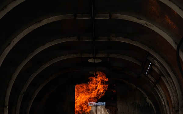The image shows the interior of a metal-walled corridor or tunnel structure with an arched ceiling, through which intense open flames are visible in the middle and background sections. The fire appears to be burning actively through a doorway or partition opening, with orange and yellow flames reaching upward toward the ceiling. The setting appears to be a controlled fire training environment or industrial facility, given the structured metal construction and compartmentalized layout.