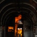 The image shows the interior of a metal-walled corridor or tunnel structure with an arched ceiling, through which intense open flames are visible in the middle and background sections. The fire appears to be burning actively through a doorway or partition opening, with orange and yellow flames reaching upward toward the ceiling. The setting appears to be a controlled fire training environment or industrial facility, given the structured metal construction and compartmentalized layout.