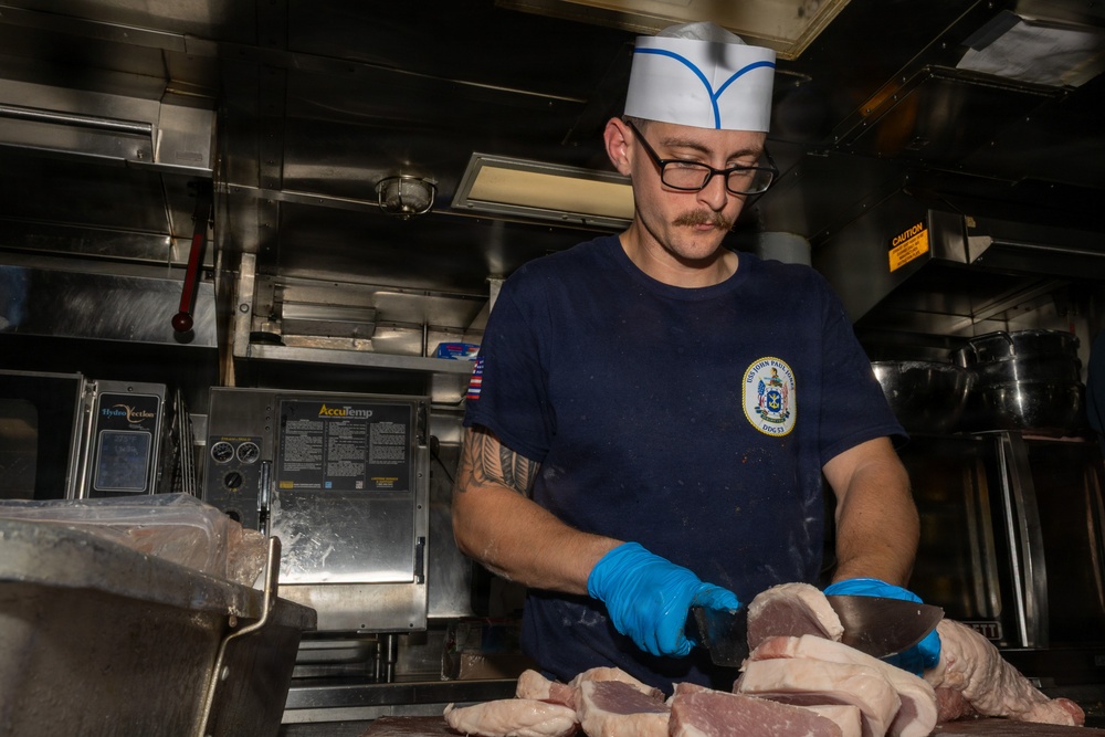 A U.S. Navy sailor wearing a white cook's hat and navy blue uniform shirt is preparing meat in a shipboard galley kitchen. The shirt patch indicates assignment to USS John Paul Jones (DDG-53). The sailor is cutting raw meat on a work surface while wearing blue nitrile gloves.