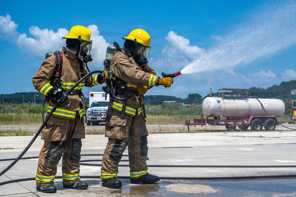 Two firefighters in full protective gear are conducting what appears to be a firefighting training exercise on a concrete pad. Both are wearing tan turnout gear with yellow reflective stripes, yellow helmets, and SCBA (self-contained breathing apparatus) units. One firefighter is actively directing a water stream from a hose nozzle toward a target area while the other provides support.