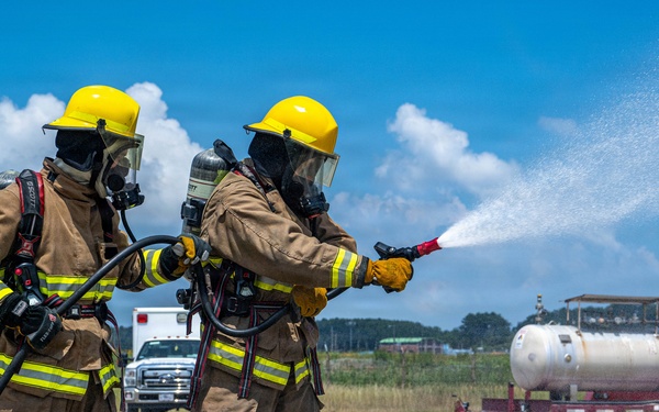 Two firefighters in full protective gear are conducting what appears to be a firefighting training exercise on a concrete pad. Both are wearing tan turnout gear with yellow reflective stripes, yellow helmets, and SCBA (self-contained breathing apparatus) units. One firefighter is actively directing a water stream from a hose nozzle toward a target area while the other provides support.