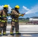 Two firefighters in full protective gear are conducting what appears to be a firefighting training exercise on a concrete pad. Both are wearing tan turnout gear with yellow reflective stripes, yellow helmets, and SCBA (self-contained breathing apparatus) units. One firefighter is actively directing a water stream from a hose nozzle toward a target area while the other provides support.