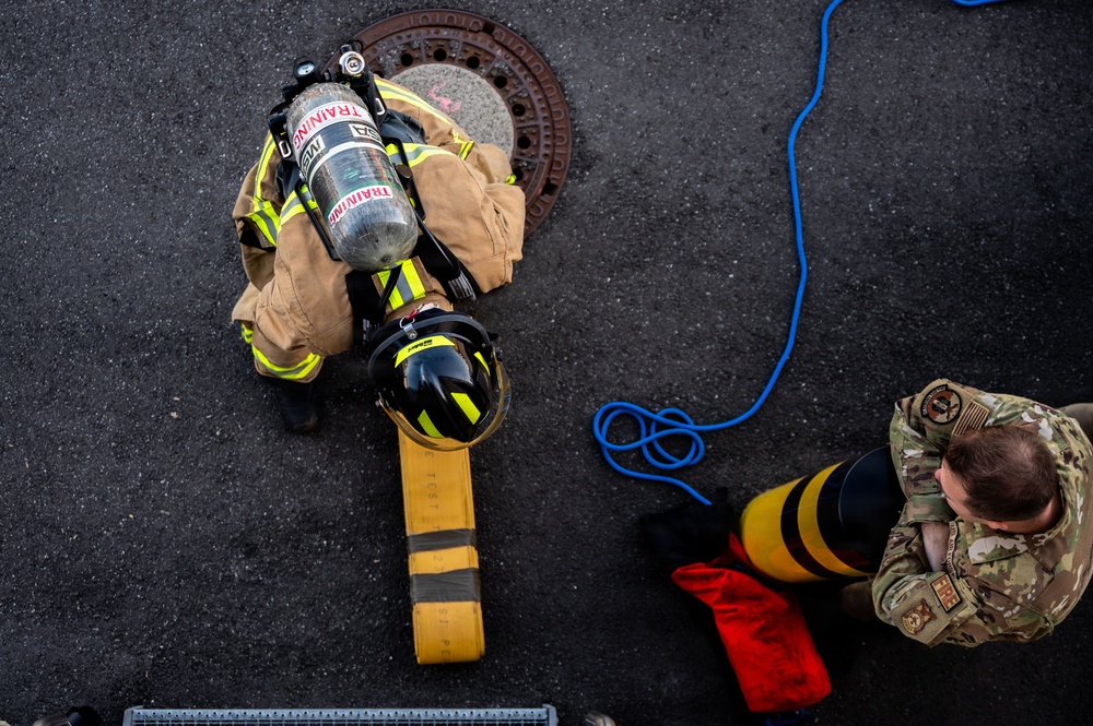 An overhead aerial view of a firefighter in full turnout gear with an SCBA (Self-Contained Breathing Apparatus) marked 'TRAINING' preparing to enter or exiting a manhole. A second individual in military camouflage (OCP/MultiCam) uniform is positioned nearby, apparently assisting or observing. The scene appears to be a confined space entry training exercise on an asphalt surface.