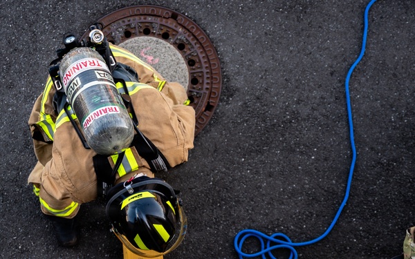 An overhead aerial view of a firefighter in full turnout gear with an SCBA (Self-Contained Breathing Apparatus) marked 'TRAINING' preparing to enter or exiting a manhole. A second individual in military camouflage (OCP/MultiCam) uniform is positioned nearby, apparently assisting or observing. The scene appears to be a confined space entry training exercise on an asphalt surface.