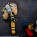 An overhead aerial view of a firefighter in full turnout gear with an SCBA (Self-Contained Breathing Apparatus) marked 'TRAINING' preparing to enter or exiting a manhole. A second individual in military camouflage (OCP/MultiCam) uniform is positioned nearby, apparently assisting or observing. The scene appears to be a confined space entry training exercise on an asphalt surface.