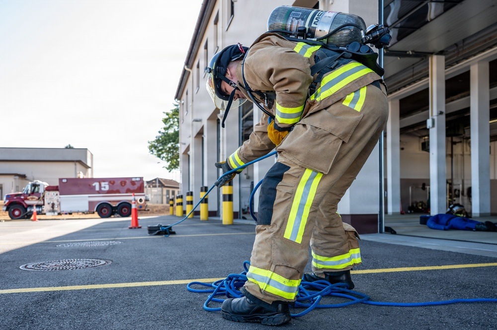 A firefighter in full personal protective equipment, including a tan turnout gear with yellow reflective stripes and an SCBA (Self-Contained Breathing Apparatus) tank, is bent over handling a blue hose near a fire station. The scene appears to be a training exercise or equipment preparation activity outside a fire station bay. A fire apparatus marked with the number '15' is visible in the background.