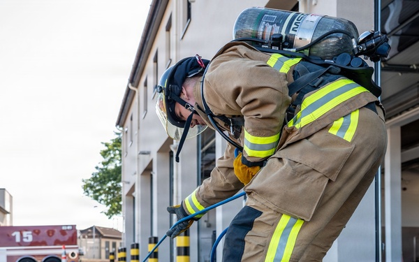 A firefighter in full personal protective equipment, including a tan turnout gear with yellow reflective stripes and an SCBA (Self-Contained Breathing Apparatus) tank, is bent over handling a blue hose near a fire station. The scene appears to be a training exercise or equipment preparation activity outside a fire station bay. A fire apparatus marked with the number '15' is visible in the background.