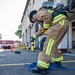 A firefighter in full personal protective equipment, including a tan turnout gear with yellow reflective stripes and an SCBA (Self-Contained Breathing Apparatus) tank, is bent over handling a blue hose near a fire station. The scene appears to be a training exercise or equipment preparation activity outside a fire station bay. A fire apparatus marked with the number '15' is visible in the background.